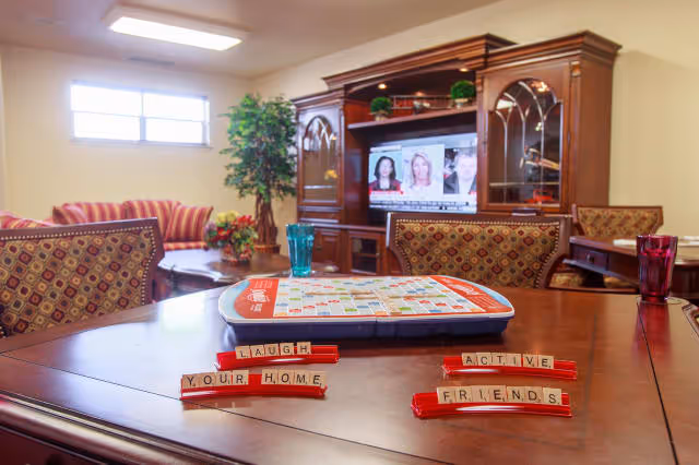 A cozy senior living common area with a wooden table in the foreground holding a Scrabble game with tiles spelling 'LAUGH', 'YOUR HOME', 'ACTIVE', and 'FRIENDS'. In the background, there is a wooden entertainment center with a TV showing a news program, a striped sofa, a potted plant, and patterned chairs around the table.