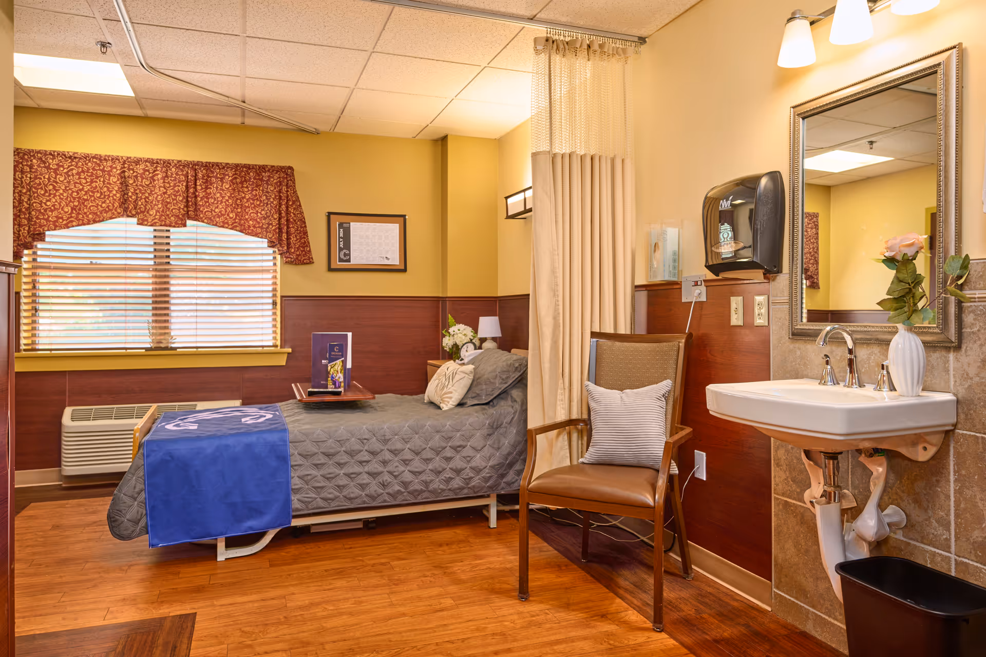 A furnished senior care bedroom with a single bed, armchair, wall-mounted sink and mirror, and a window with blinds and valance.