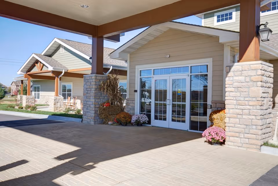 Entrance of a senior living facility with a covered driveway supported by stone pillars and wooden beams. The building has beige siding with white trim and large glass double doors. There are fall decorations including hay bales, pumpkins, and potted flowers near the entrance.