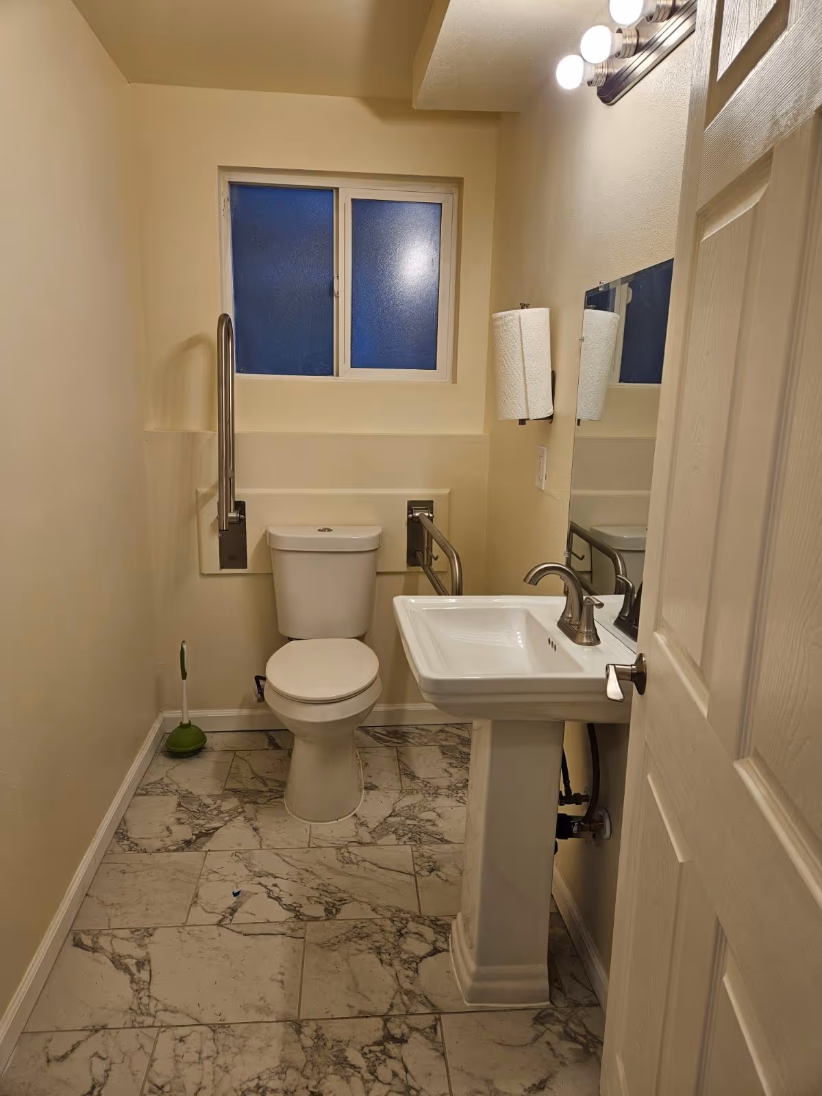Small accessible bathroom with a toilet fitted with grab bars, a pedestal sink and mirror, frosted window, and marble-patterned tile floor.