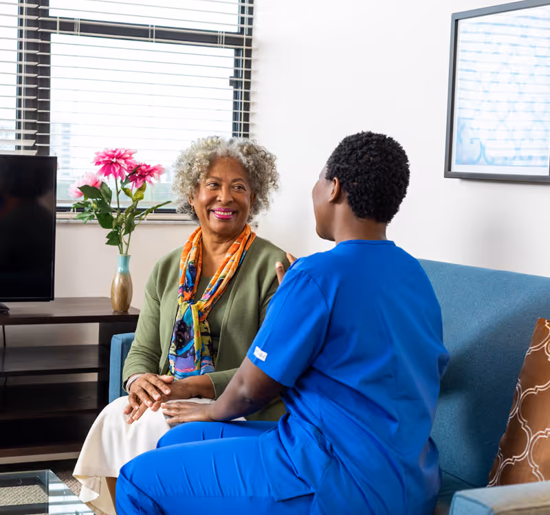 An elderly woman with curly gray hair wearing a green cardigan and colorful scarf is sitting on a blue couch, smiling and holding hands with a caregiver dressed in blue scrubs. They are in a well-lit room with a window, a vase with pink flowers on a table, a TV, and a framed picture on the wall.