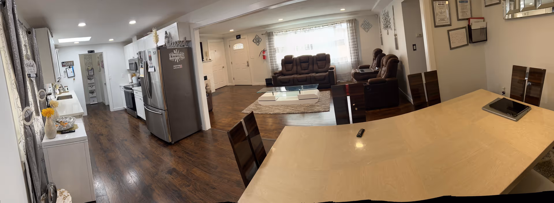Interior view of a senior living facility showing a combined kitchen, dining, and living room area. The kitchen has white cabinets, stainless steel appliances including a refrigerator and microwave, and a wooden floor. The dining area features a light wood table with dark wood chairs, a tablet, and a remote on the table. The living room has brown cushioned recliner sofas, a glass coffee table on a rug, and a large window with sheer curtains letting in natural light. The walls are decorated with framed certificates and wall art.