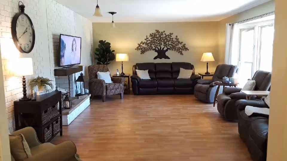 A cozy living room with wooden flooring, featuring a black leather sofa and recliners arranged around the room. There is a large wall clock on a white brick wall, a mounted flat-screen TV, two table lamps on side tables, a decorative metal tree wall art, and a large window with white curtains letting in natural light.