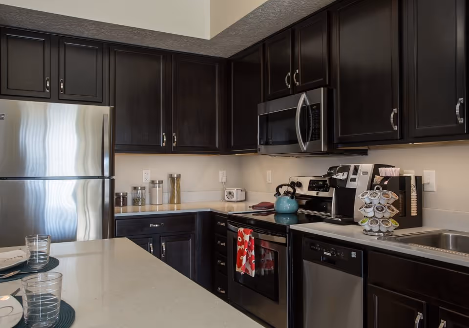 Modern kitchen with dark wood cabinets, stainless steel refrigerator, microwave, oven, dishwasher, and a coffee maker with a rotating pod holder. There is a teal kettle on the stove and a kitchen island with place settings including plates, glasses, and napkins.