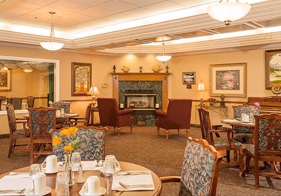 A cozy dining area in a senior living facility with round tables set with white cups, glasses, and napkins. The room features patterned upholstered chairs, two maroon armchairs facing a fireplace with a green marble surround, and framed artwork on beige walls. Soft lighting comes from ceiling fixtures and table lamps.