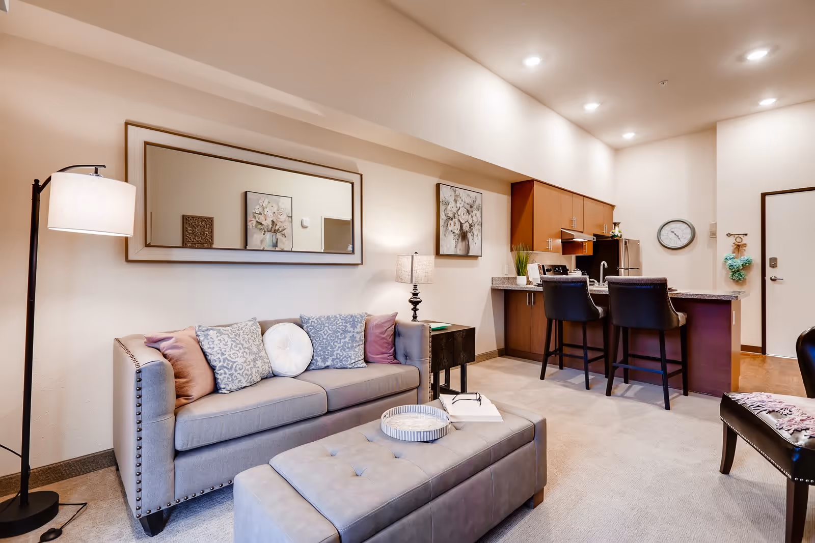 A cozy living room area with a gray sofa adorned with decorative pillows, a matching ottoman with a tray and book on top, a floor lamp, and a side table with a lamp. In the background, there is a kitchen area with wooden cabinets, a countertop with two black bar stools, a refrigerator, and a wall clock. The walls are decorated with framed floral artwork and a large mirror.