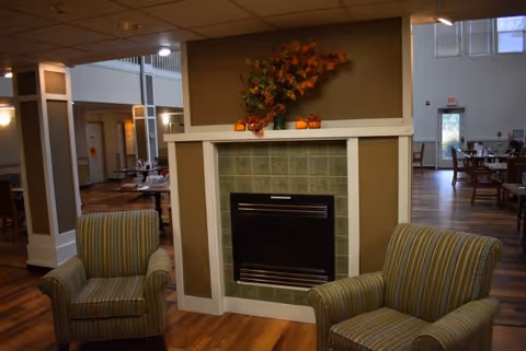 A cozy interior common area featuring a fireplace with green tiles and a white mantel decorated with autumn leaves and small pumpkins. Two striped armchairs are positioned in front of the fireplace. In the background, there are tables and chairs arranged in a dining area with large windows letting in natural light.