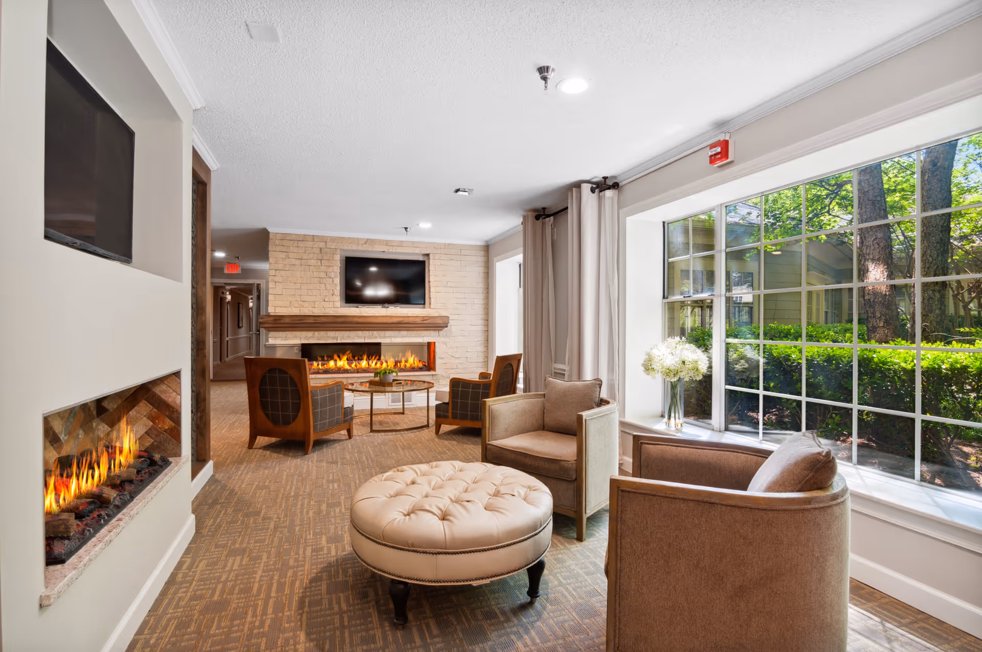 A cozy living room area with a large window showing greenery outside. The room features a modern fireplace on the left wall, a second fireplace with a mounted TV above it on the far wall, two armchairs, a round tufted ottoman, and a small table with a vase of white flowers near the window.
