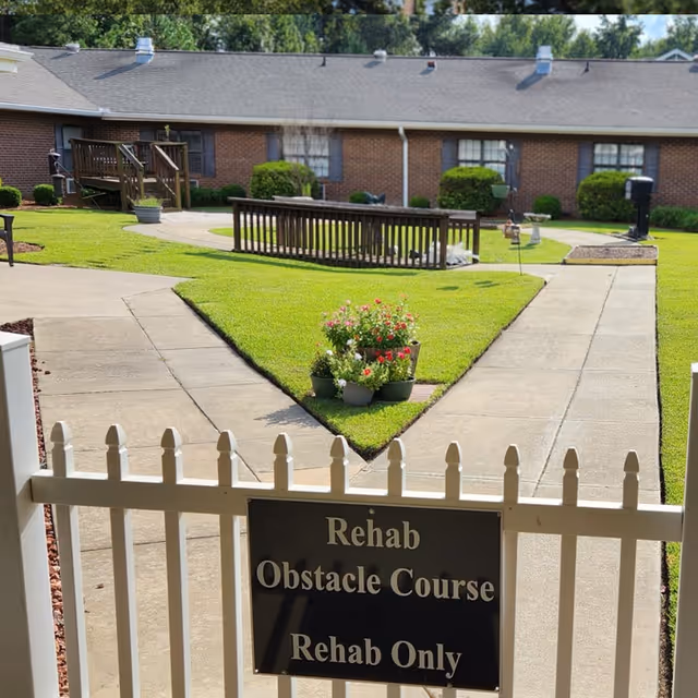 Outdoor courtyard area at Goldsboro Rehabilitation and Healthcare Center featuring a paved walkway, green grass, flower pots with colorful flowers, a wooden ramp, and a sign on a white picket fence that reads 'Rehab Obstacle Course Rehab Only'.