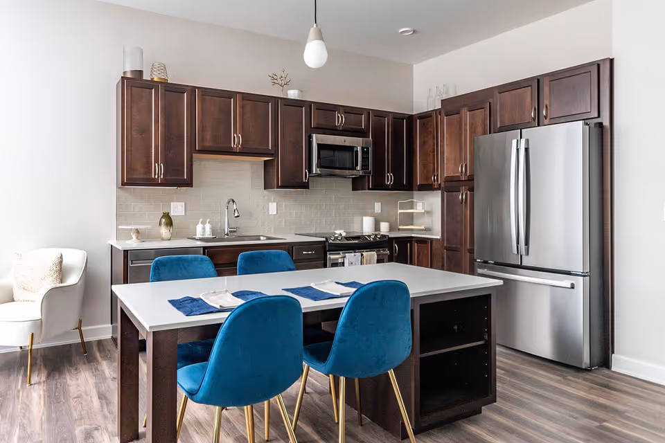Modern kitchen with dark wood cabinets, stainless steel refrigerator and microwave, a white island with blue velvet chairs, and a white armchair with a cushion in the corner.