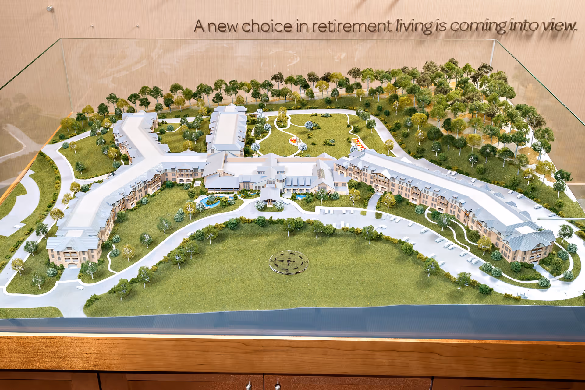 A detailed architectural scale model of a retirement living facility showing multiple buildings surrounded by green lawns, trees, pathways, and parking areas, enclosed in a glass case with the phrase 'A new choice in retirement living is coming into view' displayed above.