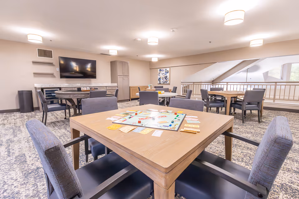 A bright, spacious common area with multiple tables and chairs arranged for social activities. One table in the foreground has a Monopoly board game set up. The room features a large flat-screen TV mounted on the wall, cabinets, and modern ceiling lights. The carpet has a patterned design, and there is a railing along one side overlooking a lower level.
