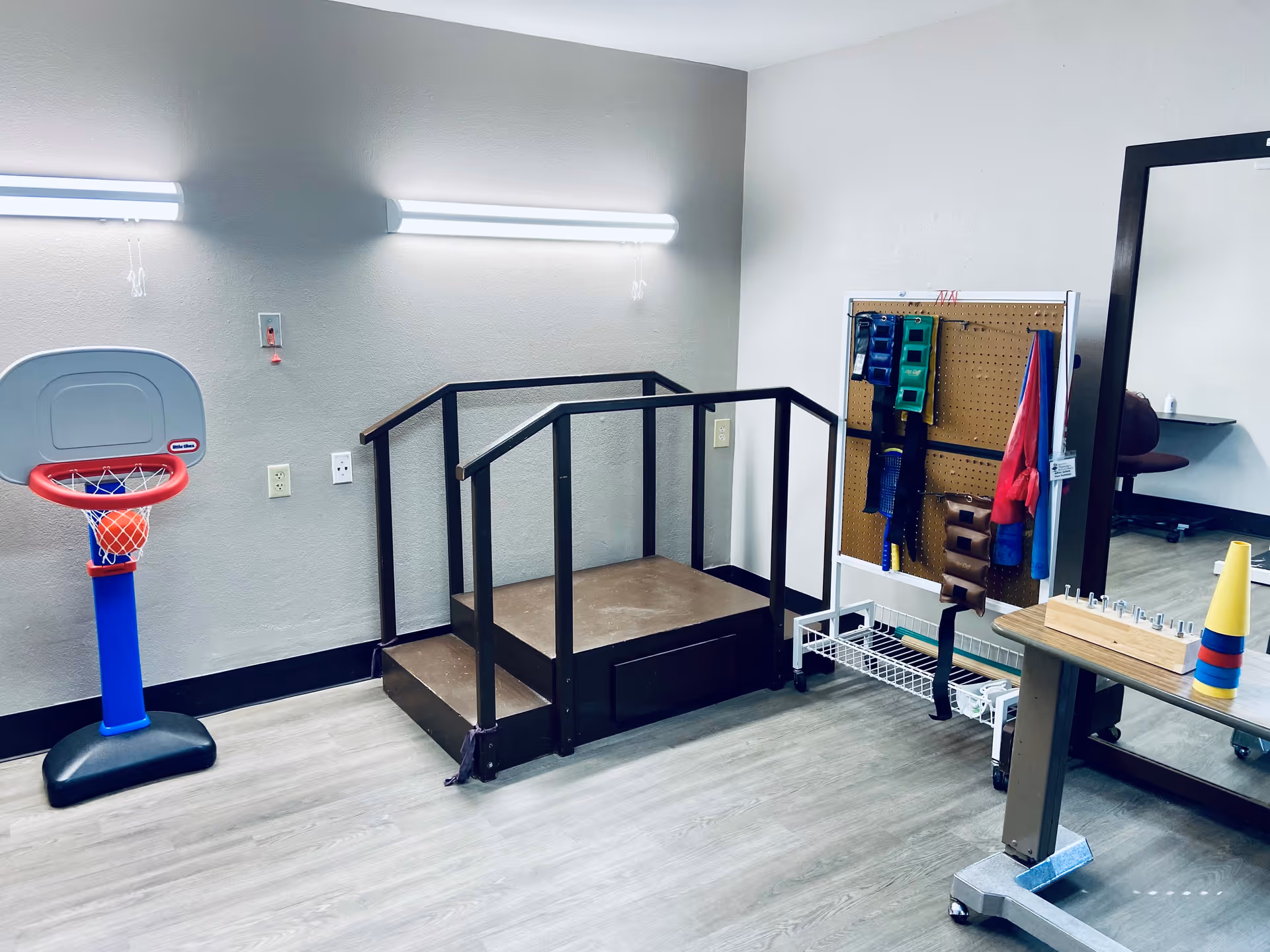 A therapy or rehabilitation room with a small basketball hoop, a wooden step platform with handrails, a pegboard holding various exercise and therapy equipment, and a large mirror on the wall. The room has light-colored walls and wood flooring.