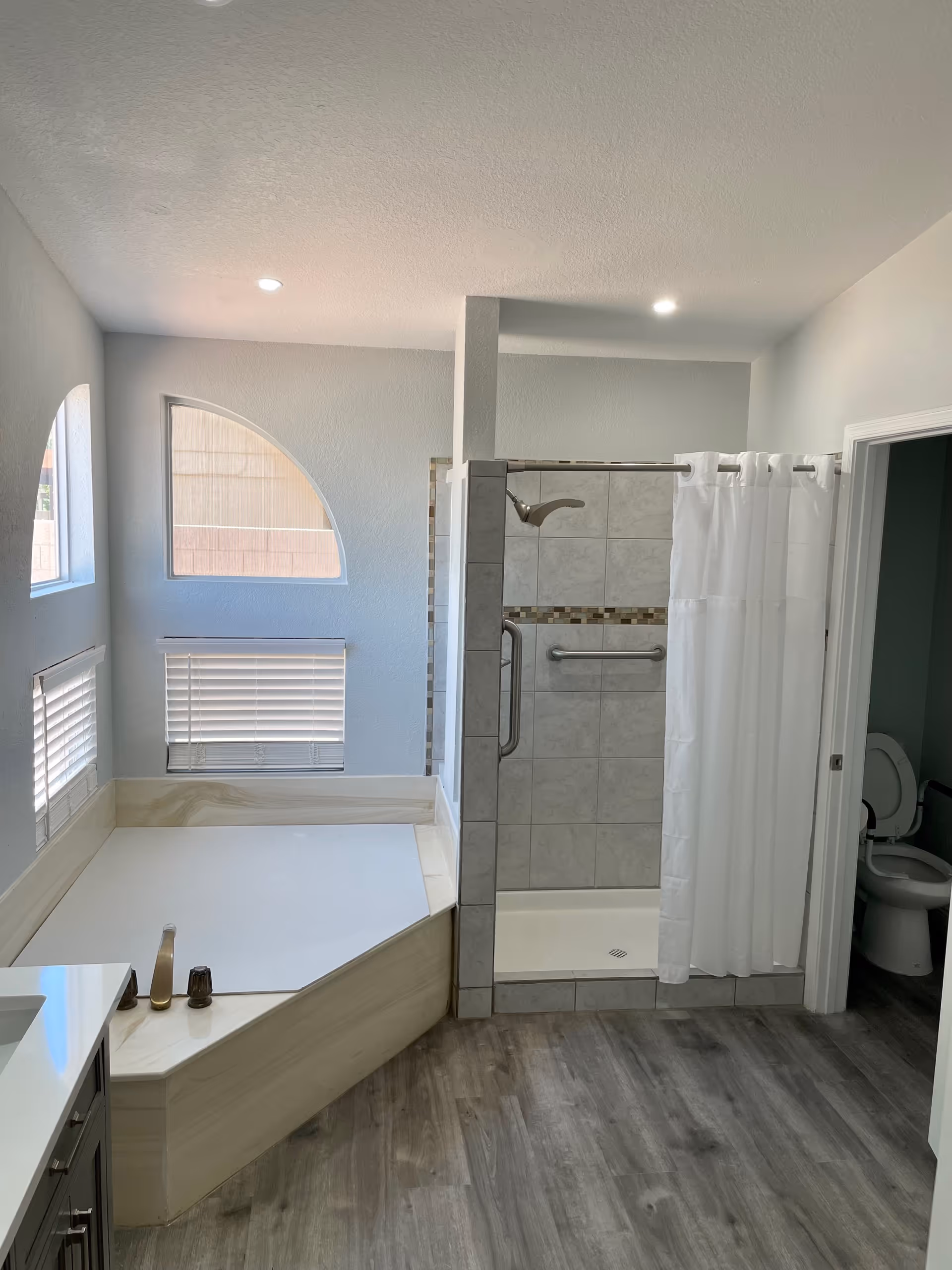 A modern bathroom with a large bathtub surrounded by beige marble, a tiled walk-in shower with a white curtain and grab bars, and a toilet visible through an open door. The room has light gray walls, wood-look flooring, and three windows with blinds allowing natural light to enter.