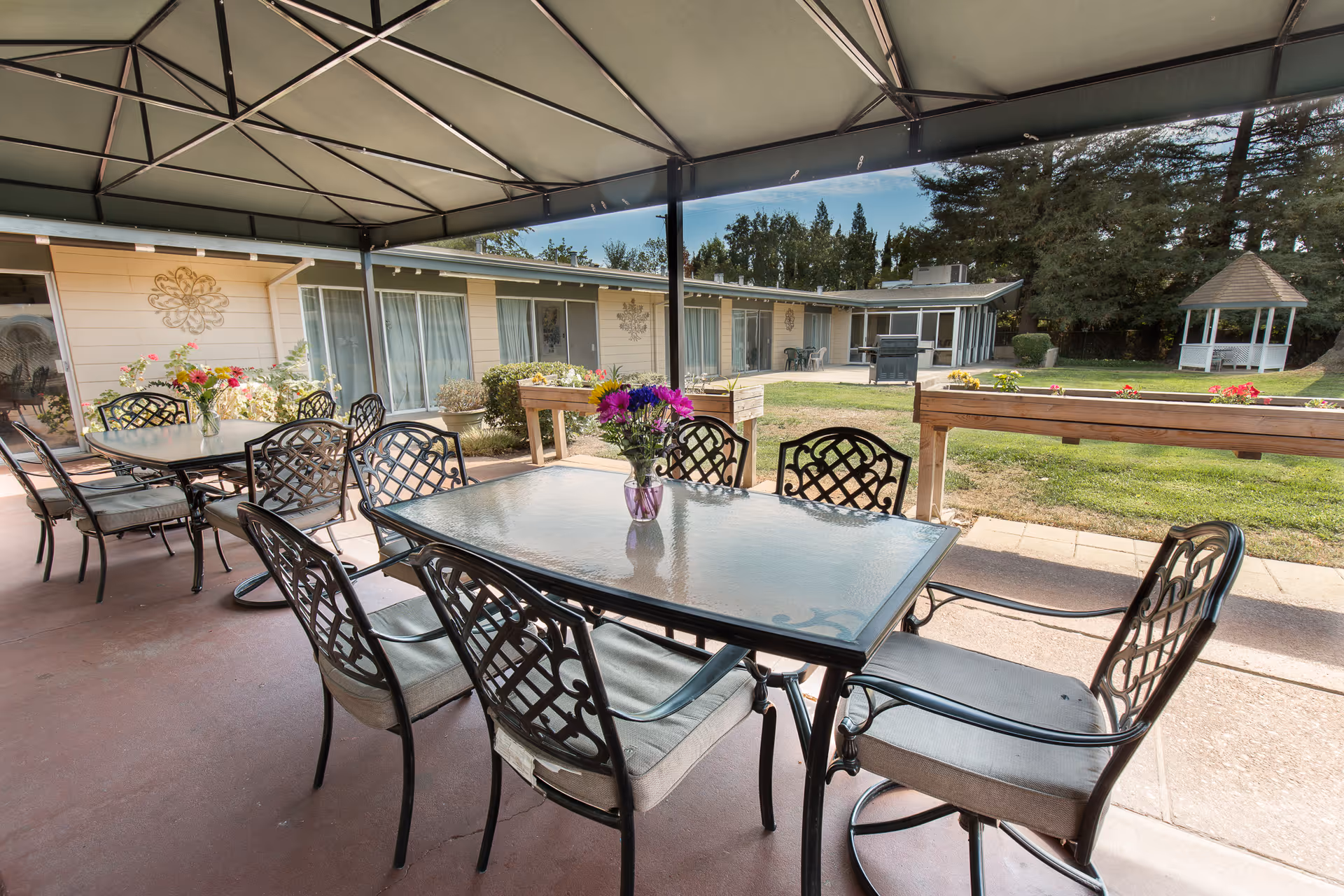 Outdoor covered patio area with glass-top tables and cushioned metal chairs. Each table has a vase with colorful flowers. In the background, there is a single-story building with large windows and sliding doors, a grassy lawn, flower beds, and a white gazebo surrounded by trees.