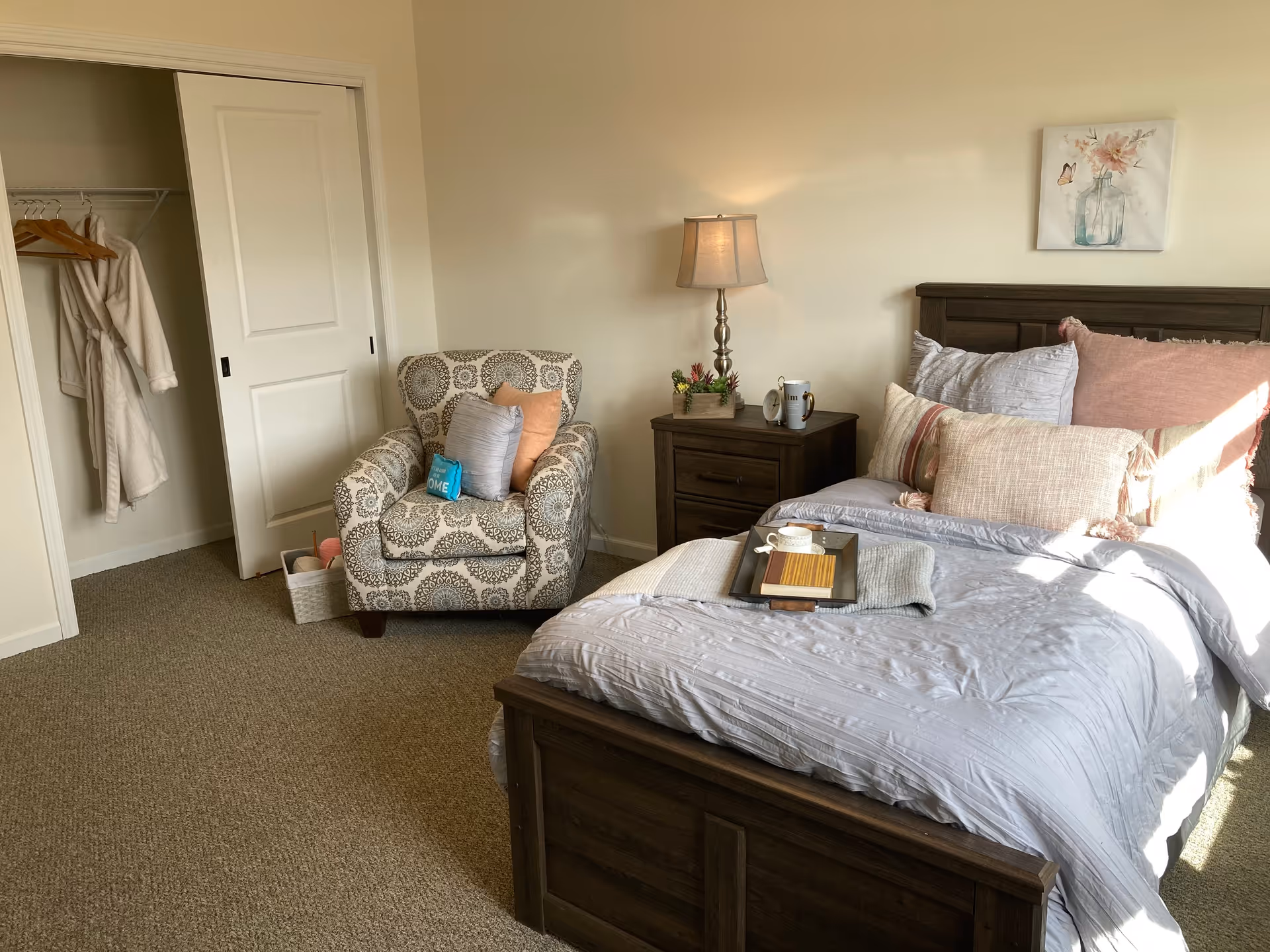 A cozy bedroom with a wooden bed frame and a neatly made bed with multiple pillows in shades of white, pink, and beige. A tray with a cup and a book rests on the bed. Next to the bed is a wooden nightstand with a lamp, a small clock, a mug, and a small plant. To the left, there is a patterned armchair with cushions and a small basket on the floor. An open closet with sliding doors reveals hanging robes and hangers. A floral painting hangs on the wall above the bed.
