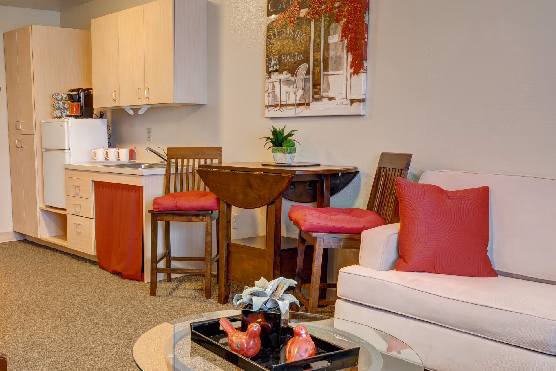 A cozy living area with a small kitchenette featuring a refrigerator, sink, and cabinets. There is a round wooden table with two chairs that have red cushions. A beige sofa with a red pillow is next to the table. A glass coffee table in front of the sofa holds a black tray with two red bird figurines and a small plant. A framed painting hangs on the wall above the table.
