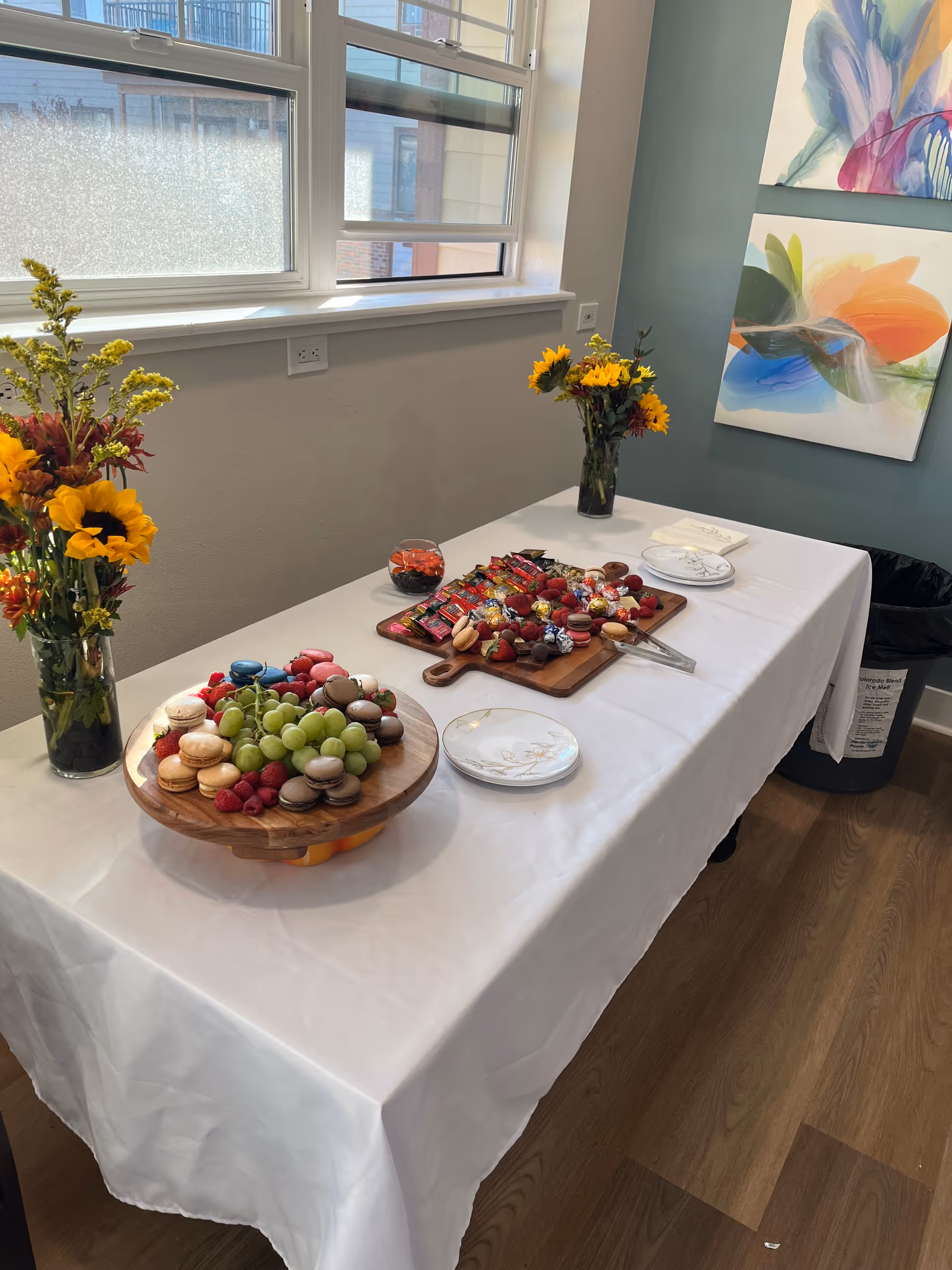 A table covered with a white tablecloth set up near a window with two vases of yellow and red flowers. On the table are two wooden platters, one with assorted macarons, grapes, and raspberries, and the other with various chocolates and candies. There are also small plates and napkins on the table. The room has light-colored walls with two colorful abstract paintings and a wooden floor.