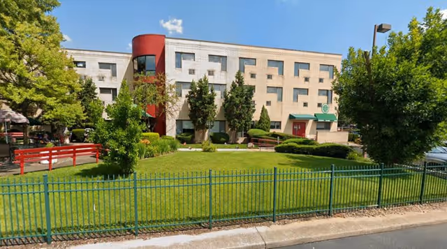 Exterior view of a three-story retirement home building with a beige facade and a red cylindrical architectural feature. The building is surrounded by green grass, trees, and bushes, with a green metal fence in the foreground. There is a red wooden bridge on the left side and a parking lot partially visible on the right. The sky is clear and blue.