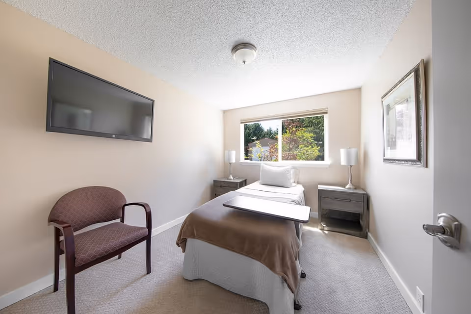 A small, bright bedroom with a single bed covered with a brown blanket and white pillow. There is a wooden overbed table on the bed, two matching nightstands with lamps on either side of the bed, a framed picture on the wall, a maroon upholstered chair, and a flat-screen TV mounted on the wall. A window shows greenery outside.