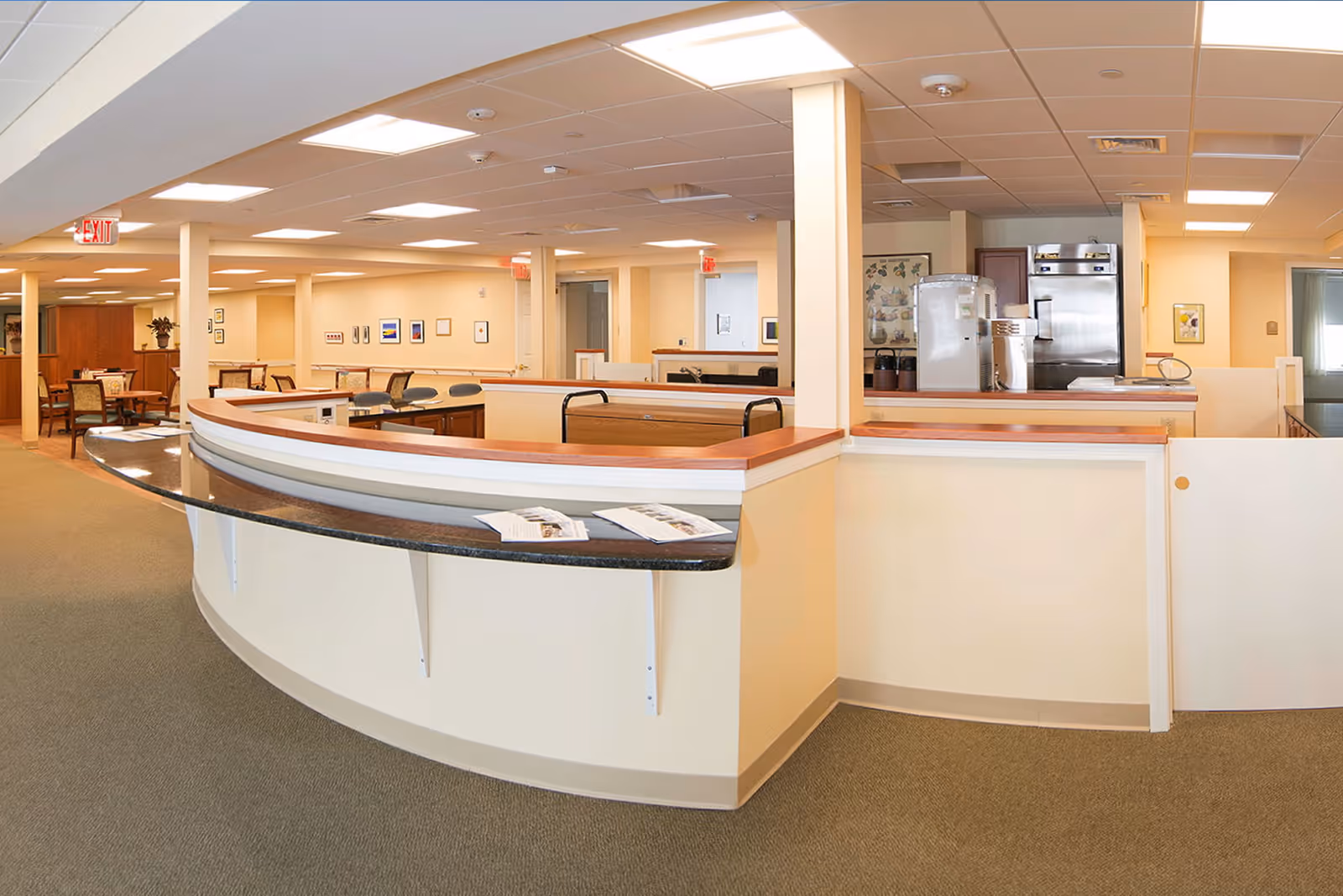 Open, well-lit reception/common area with a curved service desk, seating, and a small kitchenette in the background.