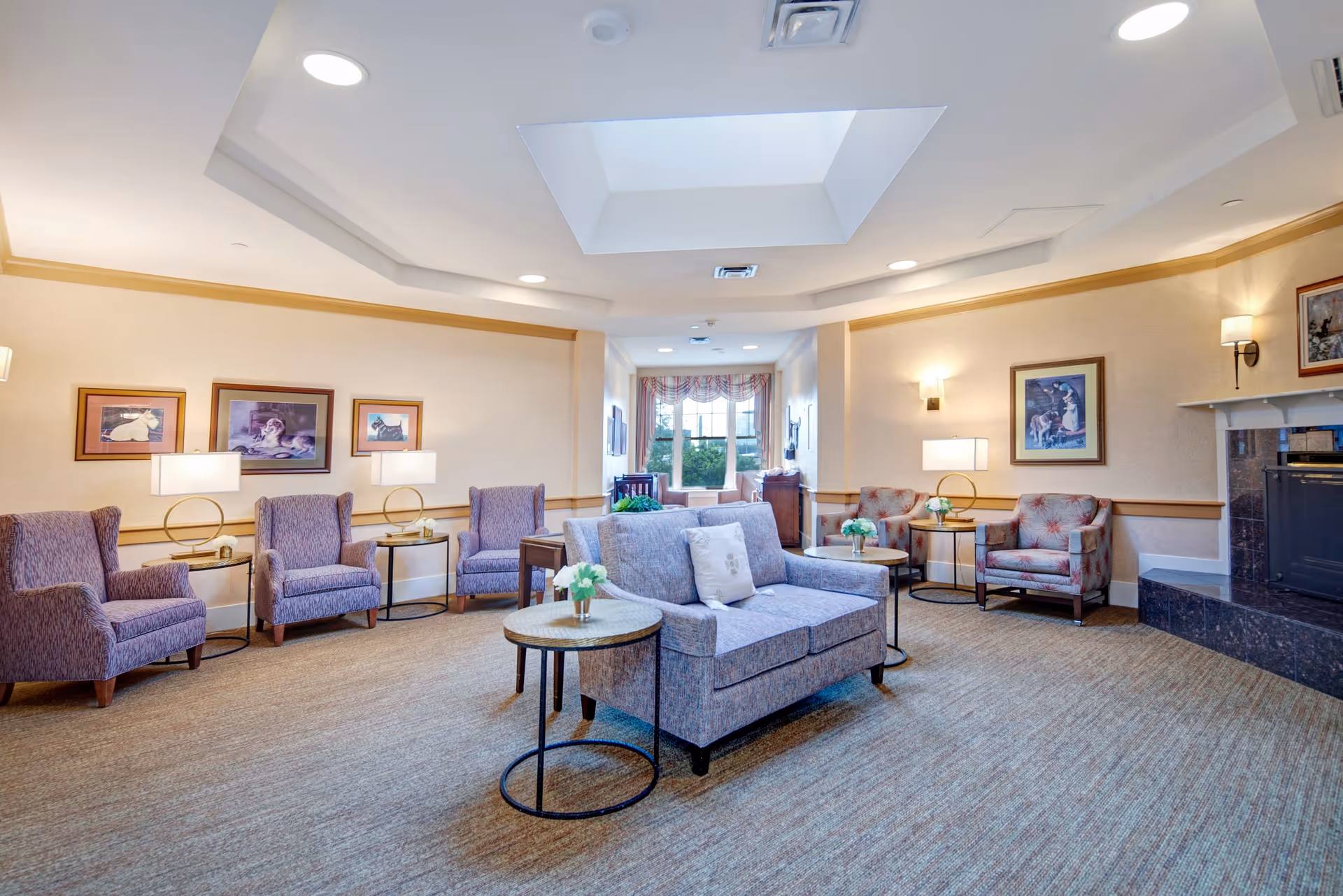 A spacious and well-lit living room with multiple upholstered armchairs and a loveseat arranged around small round tables with flower vases. The room features a skylight, framed artwork on the walls, table lamps, and a fireplace with a dark marble surround. A window with curtains is visible at the far end of the room.