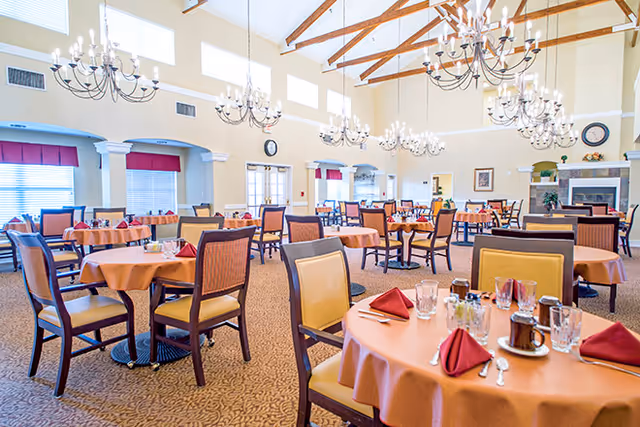 Spacious senior living dining room with round tables set with orange tablecloths and red napkins, chandeliers, and a high beamed ceiling.