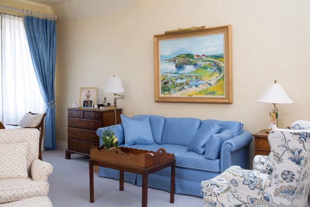 A cozy living room with a blue sofa adorned with matching blue pillows, a wooden coffee table with a small plant, a floral patterned armchair, and a beige armchair. The room features a wooden chest of drawers with framed photos and a lamp, blue curtains on a window, and a framed painting of a coastal scene on the wall.