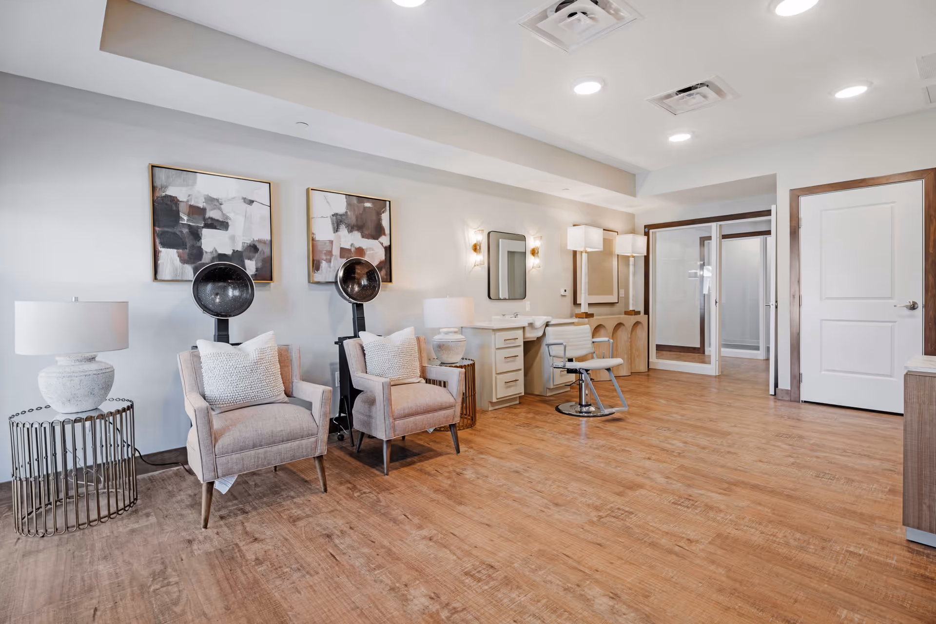 Interior view of a senior living facility hair salon area with two beige armchairs, two vintage hair dryers, a styling chair in front of a mirror and sink, modern lamps, abstract wall art, and wooden flooring.
