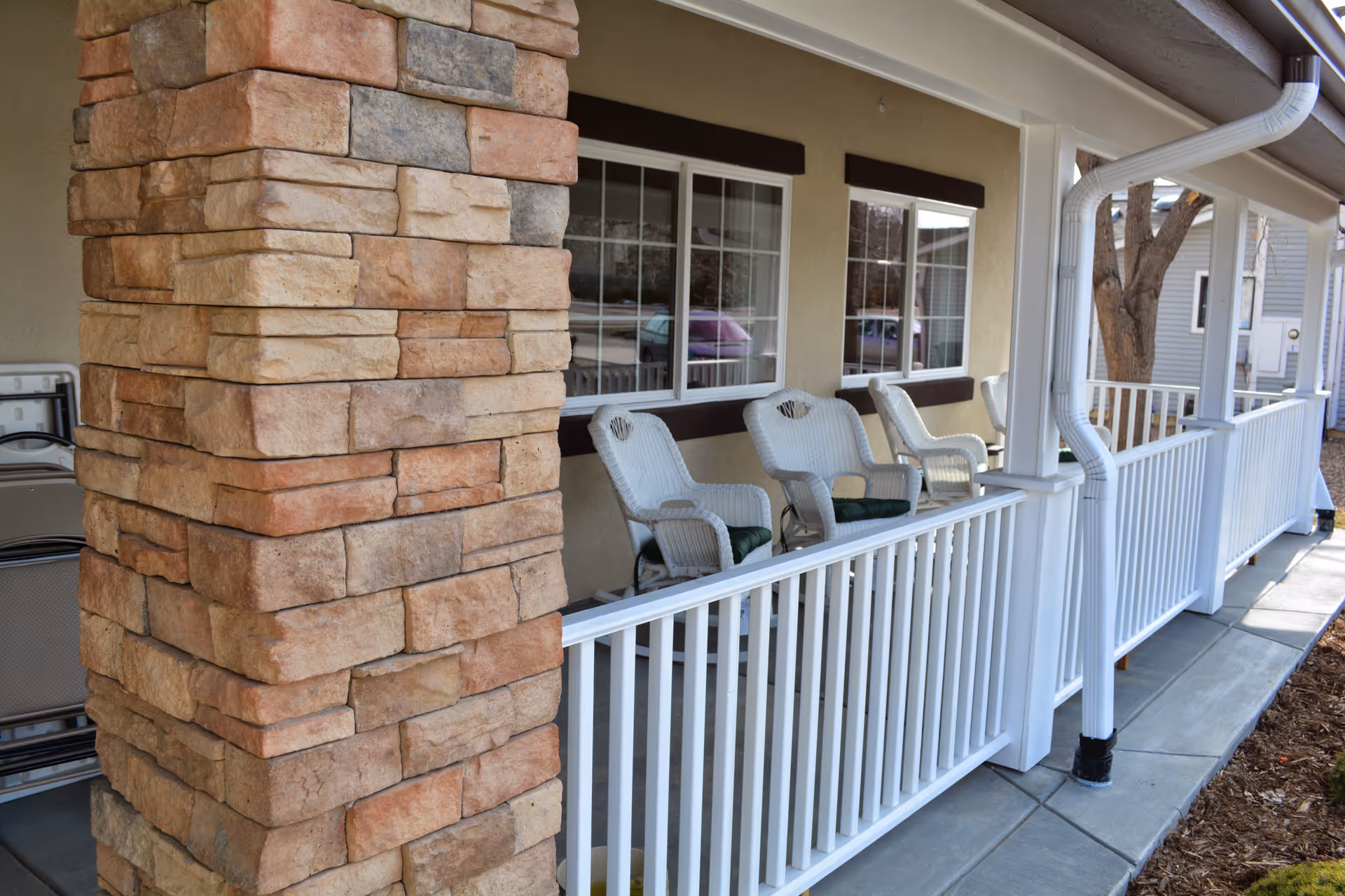 Covered front porch with a stone column, white railing, and several wicker chairs at a memory care cottage.