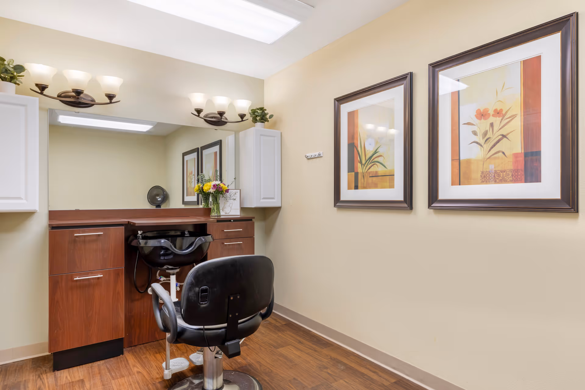 Interior view of a hair salon station in a senior living facility. The room features a black salon chair in front of a wooden counter with a built-in black sink. Above the counter is a large mirror with two wall-mounted light fixtures, each with three frosted glass shades. On the counter, there is a vase with colorful flowers and a small potted plant on a white cabinet mounted on the wall. The walls are painted beige and decorated with two framed floral artworks. The floor has a wood finish.