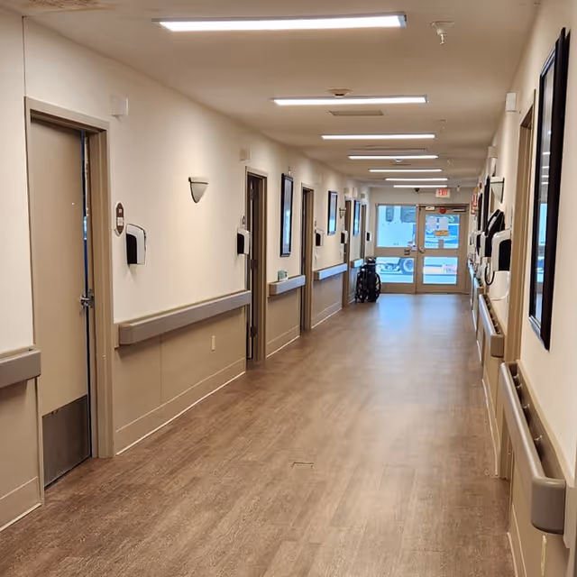 Long, well-lit hallway in a care facility with handrails along both walls, several room doors, and a wheelchair near the exit.