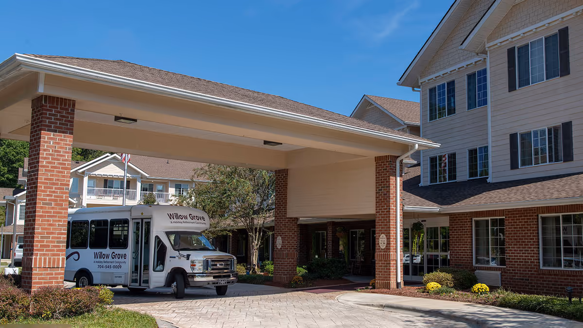Front entrance of a senior living facility with a covered porte-cochère and a 'Willow Grove' shuttle bus parked underneath.