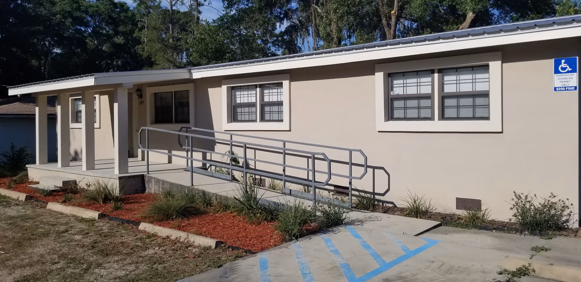 Exterior view of a single-story beige building with a covered entrance and a wheelchair accessible ramp leading to the door. There are two windows with white frames and a handicapped parking sign visible near the ramp. The surrounding area has some landscaping with small plants and red mulch.