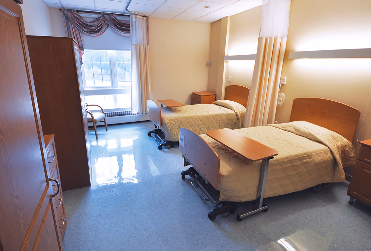 A bright and clean healthcare facility room with two single beds, each with a beige quilted bedspread and an overbed table. The room has light yellow walls, a large window with curtains, wooden furniture including dressers and nightstands, and a chair near the window. The floor is a shiny light blue linoleum.