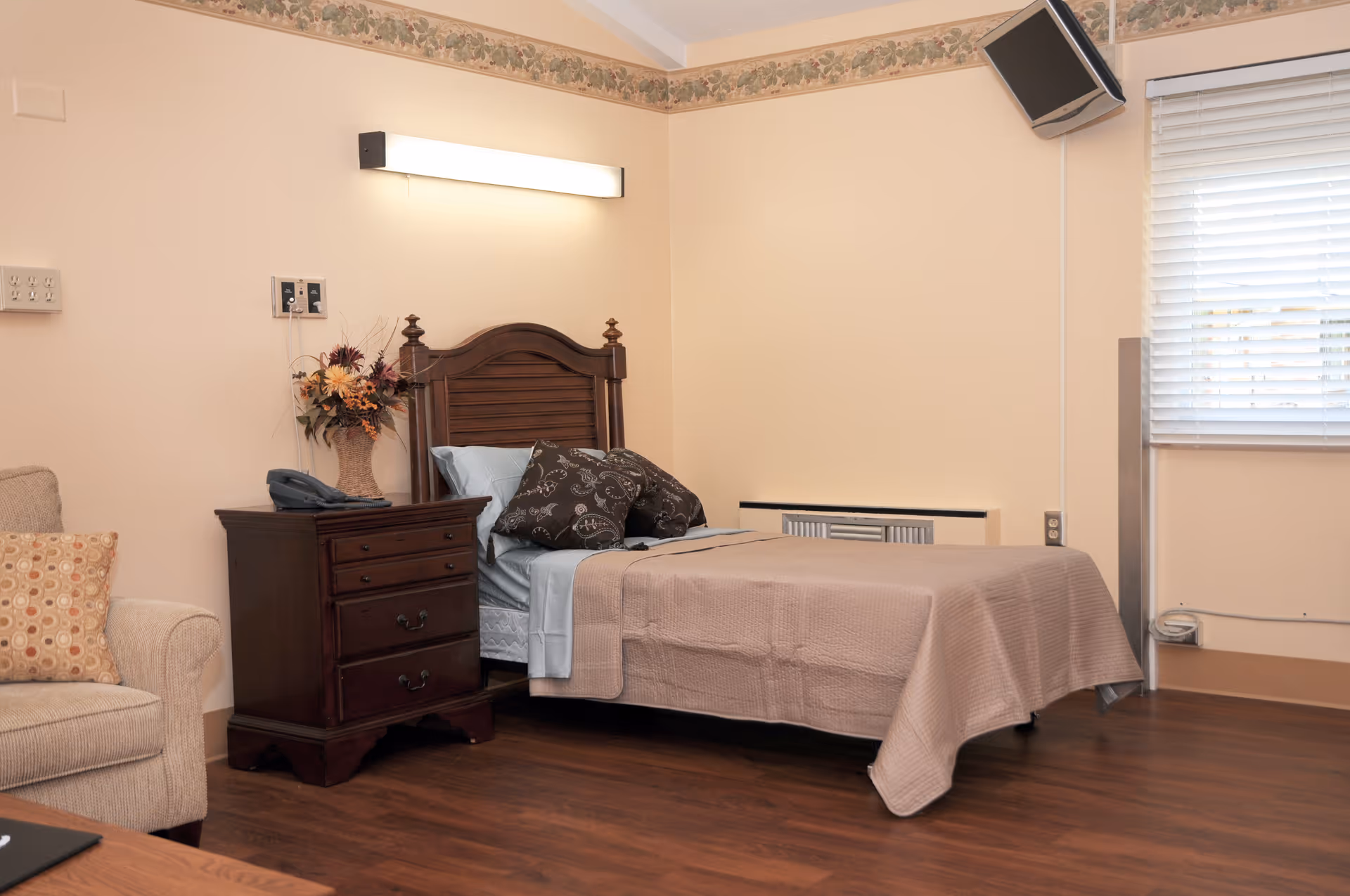 Furnished single bedroom with a wooden headboard bed, nightstand with phone and flowers, an armchair, and a wall-mounted TV.