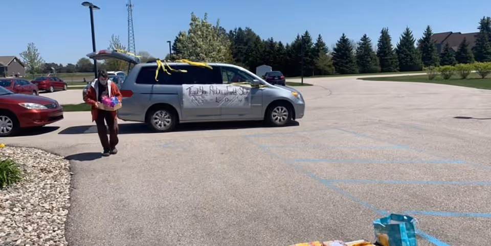 A person wearing a mask and a red jacket is walking in a parking lot carrying a box with colorful items. A gray minivan with its rear hatch open is parked nearby, decorated with yellow streamers and a large white sign on the side that reads 'Thank You Auburn Fields'. Several other cars and trees are visible in the background under a clear blue sky.