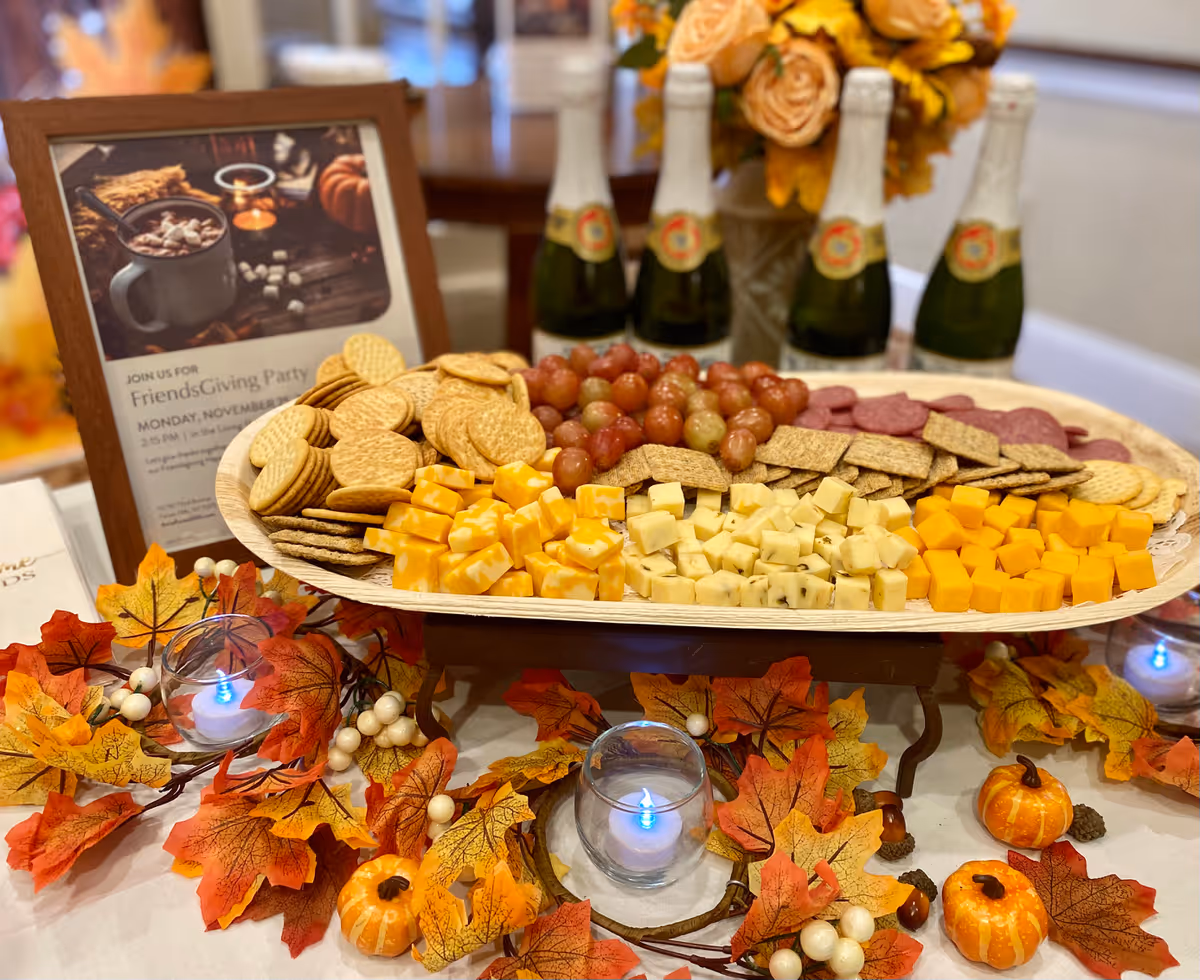 A festive platter with an assortment of crackers, cubed cheeses, grapes, and slices of salami on a table decorated with autumn leaves, small pumpkins, and white berries. Behind the platter are five bottles of sparkling cider or champagne and a framed sign advertising a FriendsGiving Party. There are also small blue LED candles in glass holders on the table.