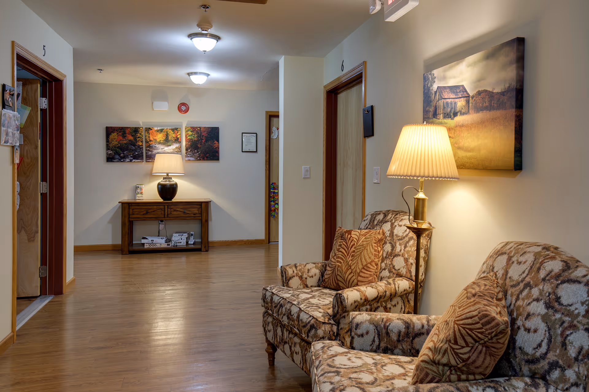 A cozy hallway in a senior living facility with two patterned armchairs and a floor lamp on the right side. The walls are decorated with framed pictures, and there is a wooden console table with a lamp and decor at the end of the hallway. Doors numbered 5 and 6 are visible along the hallway.