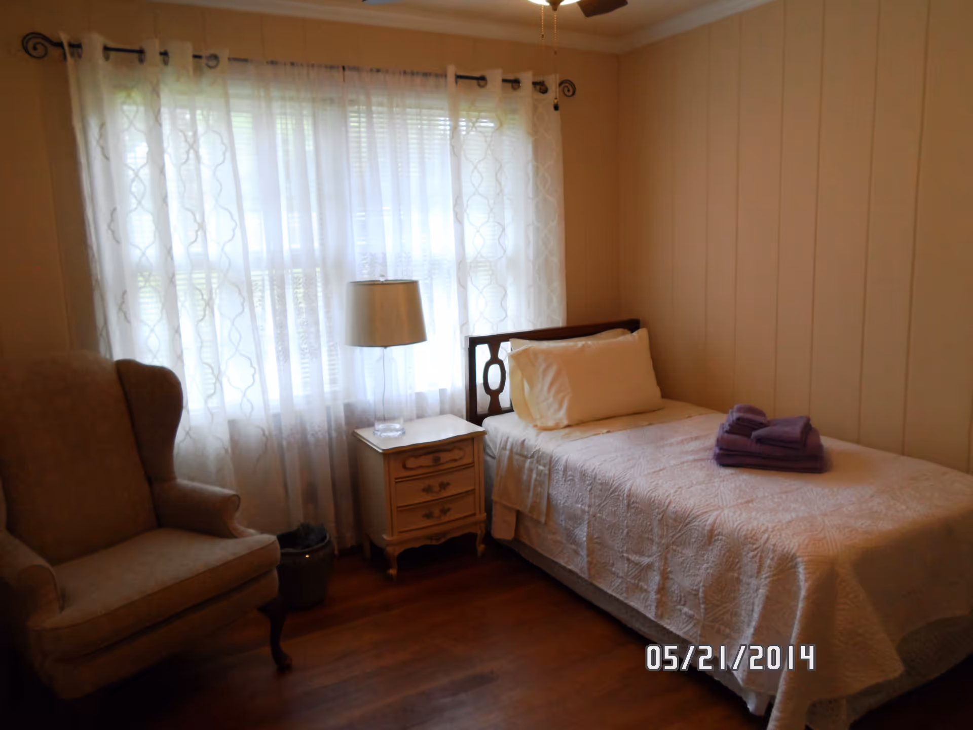 A small bedroom with a single bed covered in a white quilt and a stack of folded purple towels on top. Next to the bed is a small nightstand with a lamp. There is a beige upholstered armchair to the left of the nightstand. The room has wooden floors and light-colored walls with a window covered by sheer white curtains.