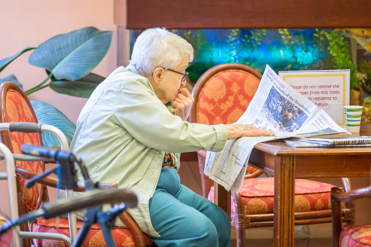An elderly woman with white hair and glasses is sitting on a red cushioned chair at a wooden table, reading a newspaper. There is a walker in the foreground and a large green plant in the background. A sign on the table requests not to remove the newspaper from the room so all residents can enjoy it.