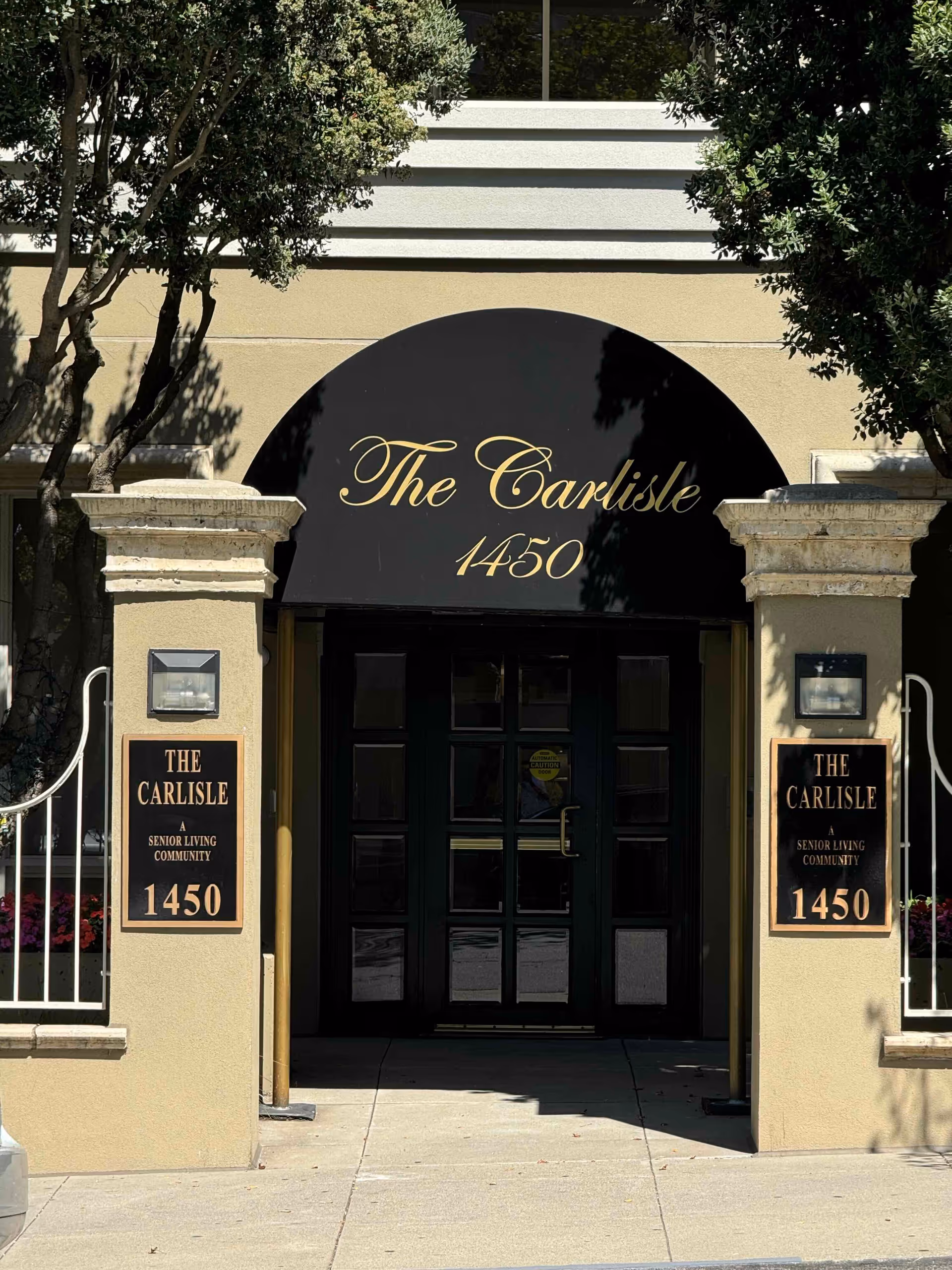Entrance of The Carlisle senior living community with a black arched awning reading "The Carlisle 1450" above double doors and pillar signs.