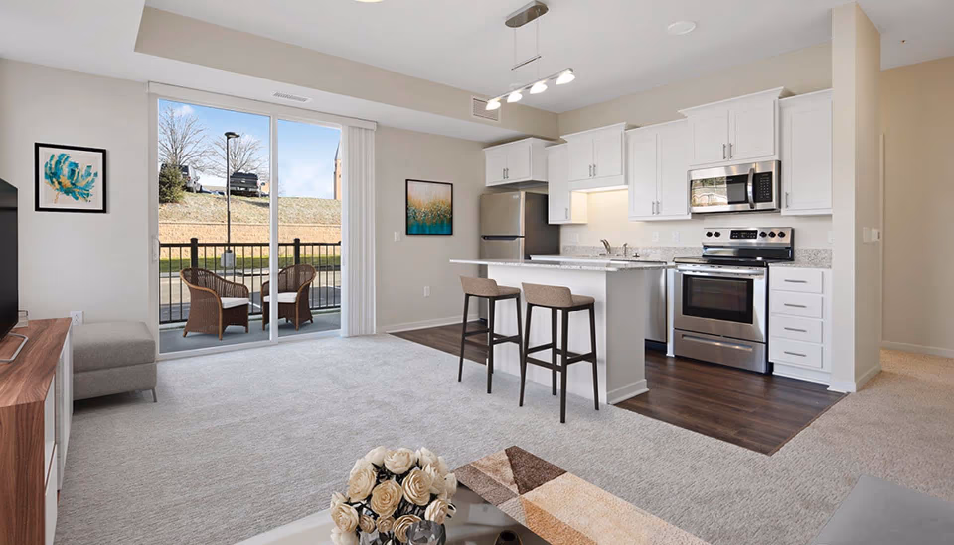 A modern open-concept living space featuring a kitchen with white cabinets, stainless steel appliances, and a breakfast bar with two stools. The room has light-colored carpet and wood flooring in the kitchen area. A sliding glass door leads to a small outdoor patio with two wicker chairs. The walls are decorated with framed artwork, and a TV stand with a television is partially visible on the left.