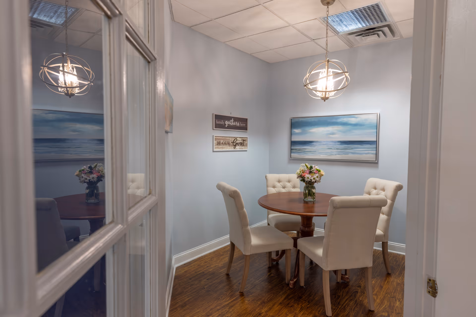 Small dining/meeting room with a round wooden table surrounded by four tufted upholstered chairs, a floral centerpiece, coastal artwork on light blue walls, and a pendant light.
