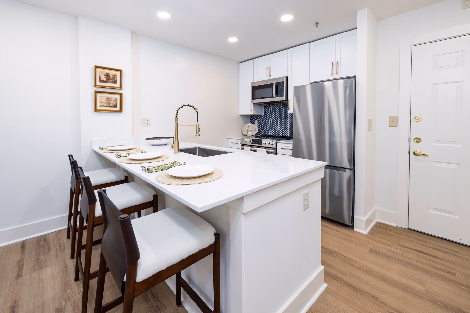 Modern kitchen with white cabinetry, stainless steel refrigerator and microwave, a white countertop island with a built-in sink and gold faucet, and three wooden bar stools with white cushions. The kitchen has a wooden floor and two framed pictures on the wall.