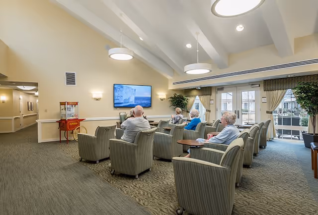 A group of elderly people seated in comfortable armchairs arranged in rows facing a wall-mounted television in a well-lit common room with beige walls and carpeted floors. There is a popcorn machine on the left side near a hallway, large windows with curtains on the right, and ceiling lights providing illumination.