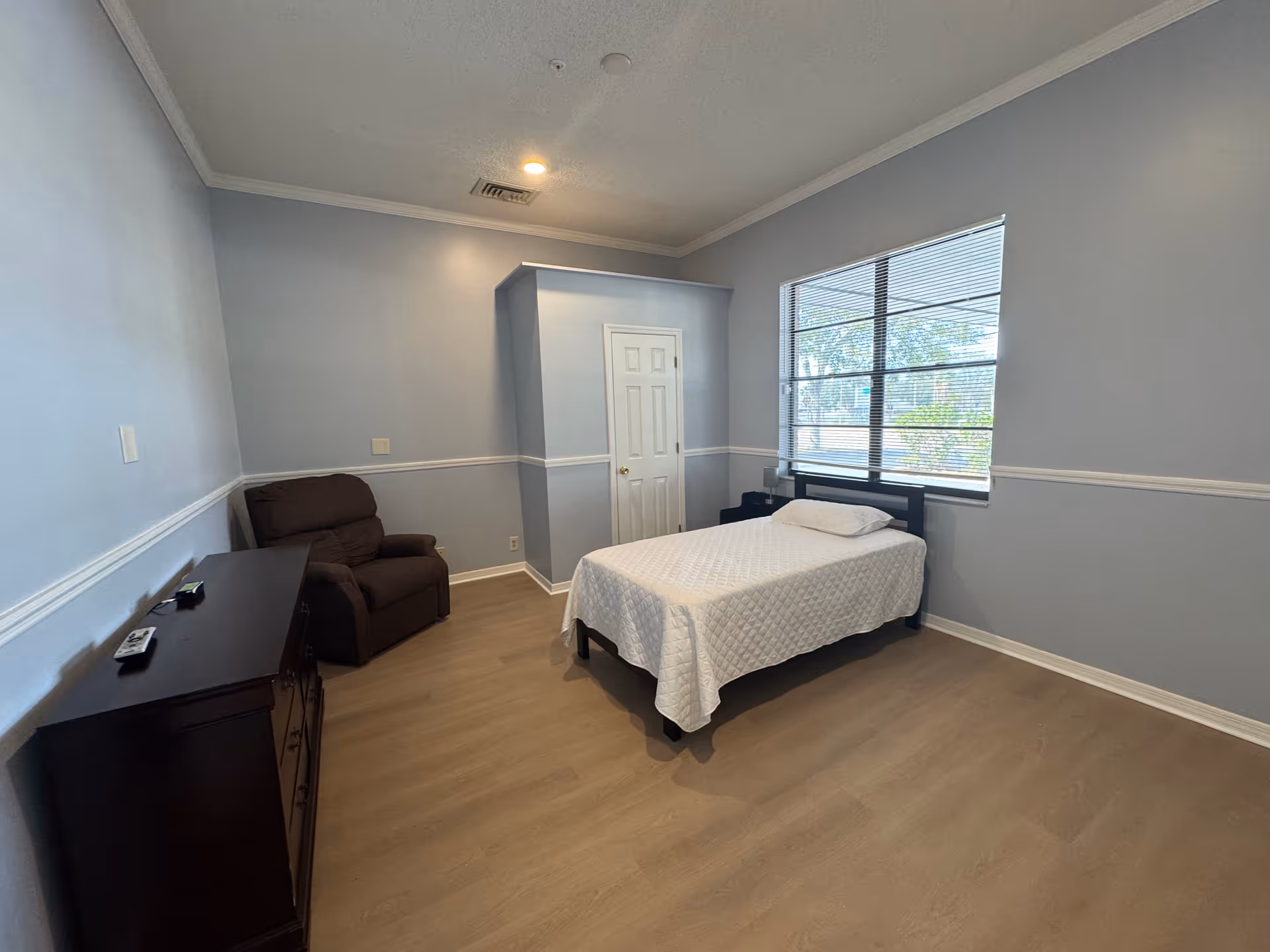 A simple bedroom with light blue walls and wood flooring. The room contains a single bed with a white quilt and pillow, a dark brown armchair, a dark wooden dresser with a remote control on top, and a window with blinds letting in natural light. There is a small closet with a white door in the corner.