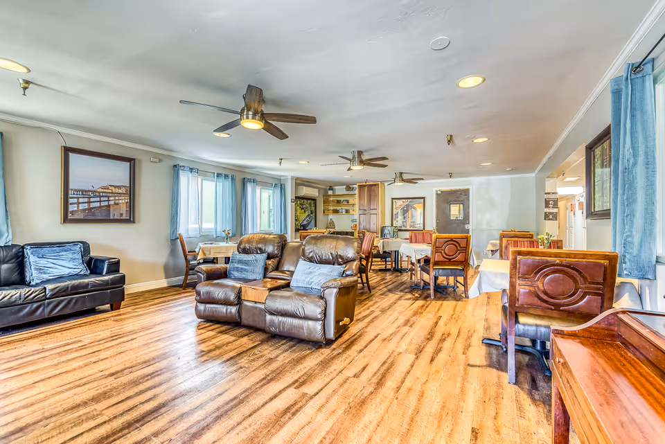 A spacious room with wooden flooring featuring multiple seating areas including a black leather couch, two brown leather recliners with blue pillows, and several wooden dining tables with chairs covered in white tablecloths. The room has ceiling fans, large windows with blue curtains, framed pictures on the walls, and a bright, welcoming atmosphere.