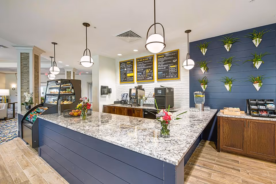 A modern cafe counter area inside a senior living facility with a granite countertop, hanging pendant lights, a menu board on a white brick wall, a display case with food items, and a blue wall decorated with small potted plants. There are flowers in vases on the counter and a water dispenser on a side cabinet.