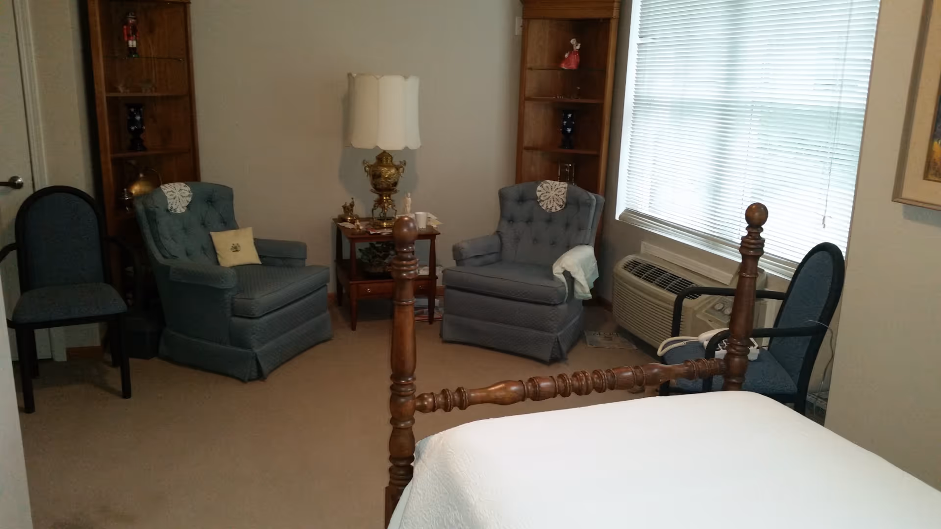 A cozy bedroom with a wooden bed frame and white bedding in the foreground. The room features two blue upholstered armchairs with doilies on the backrests, a small wooden side table with a decorative lamp and figurines, two wooden bookshelves with decorative items, and two additional blue chairs. A window with closed blinds and an air conditioning unit is on the right wall.